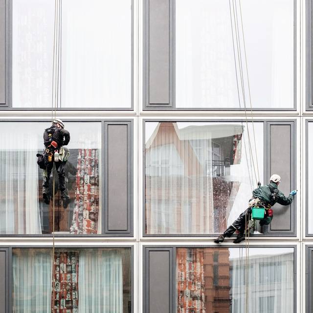 Photo d’illustration : deux laveurs de vitres en rappel sur la façade d’un immeuble aux fenêtres à cadre gris.