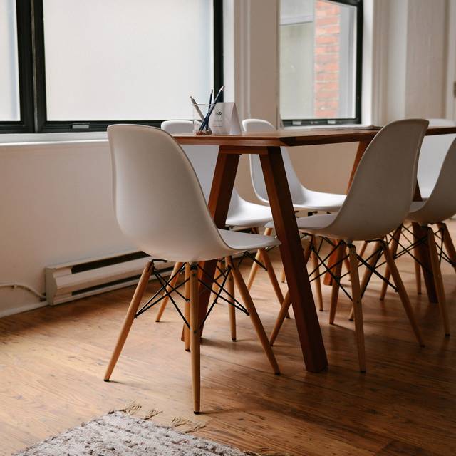 Photo d’illustration : une table en bois et des chaises blanches devant une fenêtre à store.
